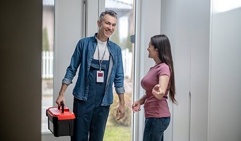 Visiteur souriant avec une boîte à outils, accueillant une résidente dans un environnement moderne et professionnel, illustrant un service d'entretien ou de dépannage à domicile.